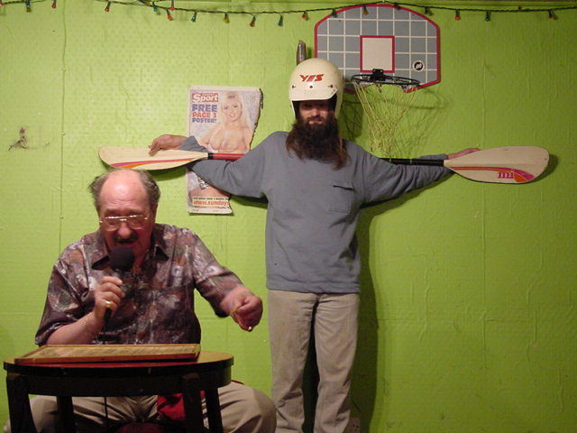 Gordon Cragg seated at table calling bingo numbers while a Jesus like figure stands behind in a crash hat on and using a canoe paddle for a cross